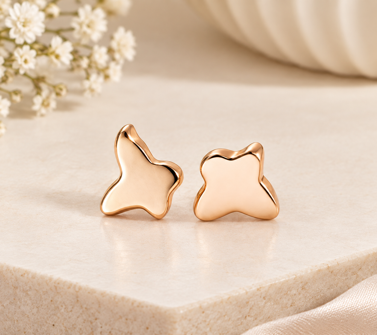 Pair of polished gold-tone butterfly-shaped stud earrings displayed on beige stone beside white flowers.