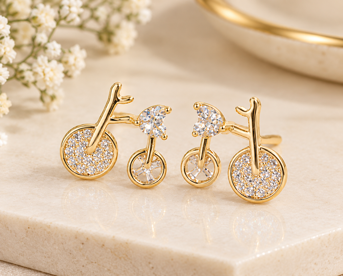 Gold-tone bicycle-shaped earrings with clear heart-shaped stones and sparkling stone-covered wheels on a beige display.