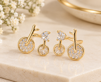 Gold-tone bicycle-shaped earrings with clear heart-shaped stones and sparkling stone-covered wheels on a beige display.