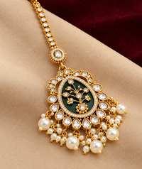 Gold-tone maang tikka with a dark enamel floral centre, clear stone border and dangling white pearl-style beads.